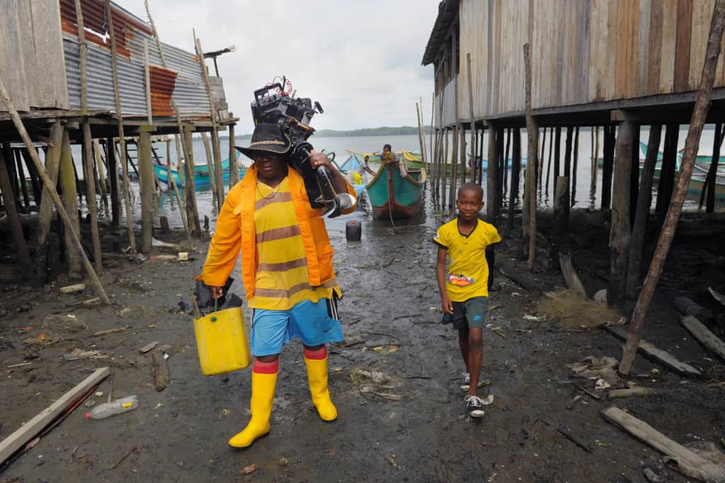 A man carries a bucket and a boat motor as he walks with a boy in from the water.