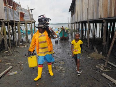 A man carries a bucket and a boat motor as he walks with a boy in from the water.