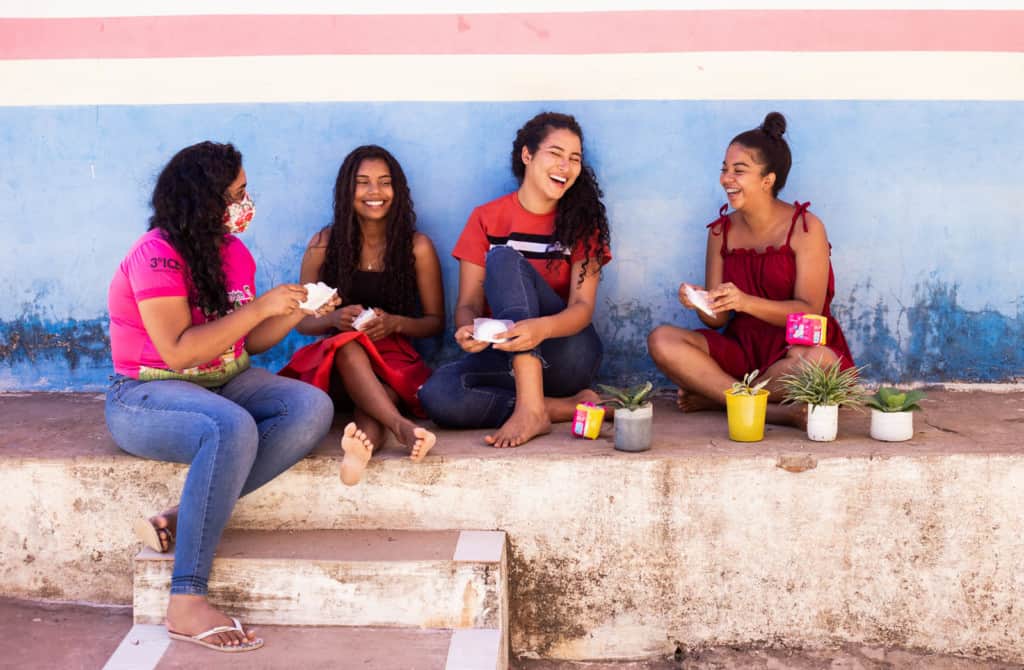 Maria Rita is wearing a red shirt and jeans. She is sitting outside the center with two of her friends and a tutor. They are talking about pads and menstruation.