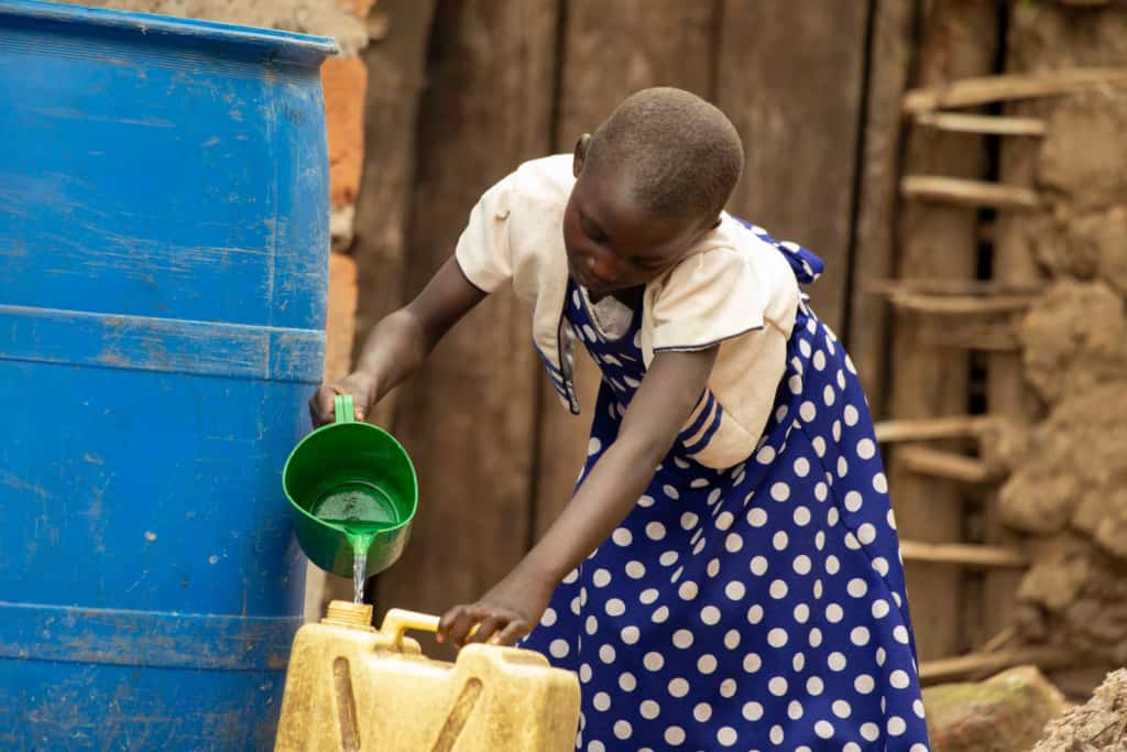 Promise is wearing a blue and white polka dotted dress with a white jacket. She is pouring water into a yellow jerry can from a large blue rainwater harvesting tank.