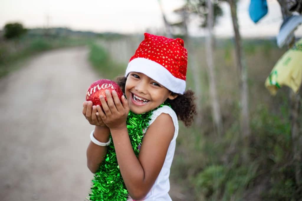 Grazielly is holding a Christmas ball that says “Merry Christmas” in Portuguese. SHe is wearing a red Santa hat and green garland around her neck. She is standing outside.