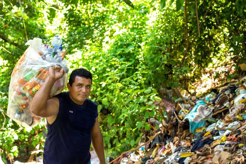 Orlando, wearing a dark blue shirt, is looking for items he can recycle in the local landfills. He is holding up a bag full of items he has found to sell.