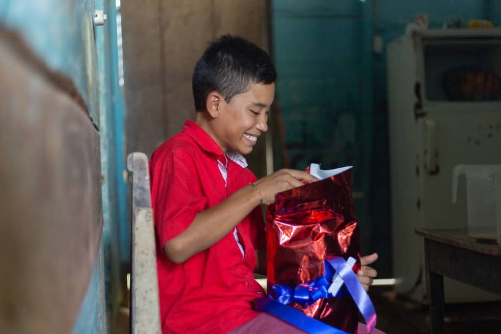 Coty is wearing a red shirt. He is sitting at home  opening his Christmas gift from the Compassion center.