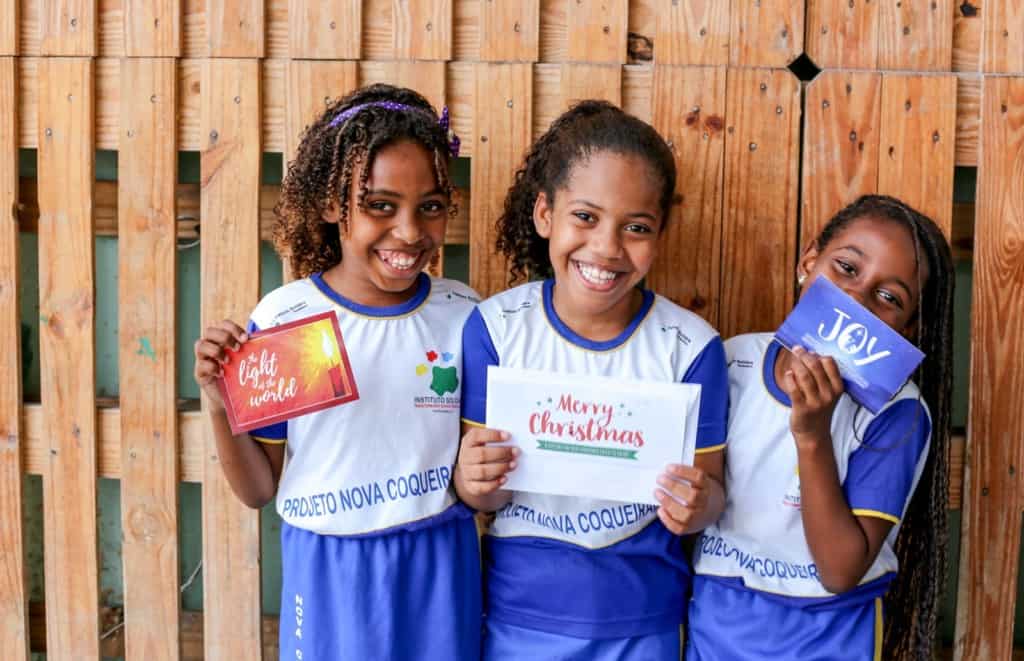 Compassion children in Brazil are joyful to receive Christmas cards from their sponsors from different parts of the world. Three smiling girls wearing blue and white clothing are holding their Christmas cards from the United States. A wooden fence is in the background. 
