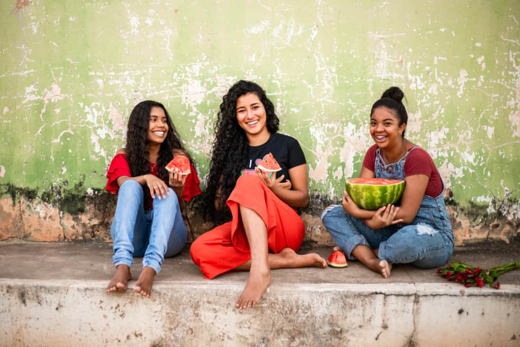 Maria Rita is wearing a red shirt and jeans. She is sitting in front of a green building with two of her friends. They are all eating watermelon.