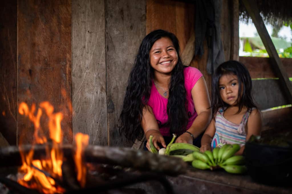 Jaela is wearing a pink shirt. She is in the kitchen of her home with her younger sister, Mayte. They are cooking plantains over an open fire.