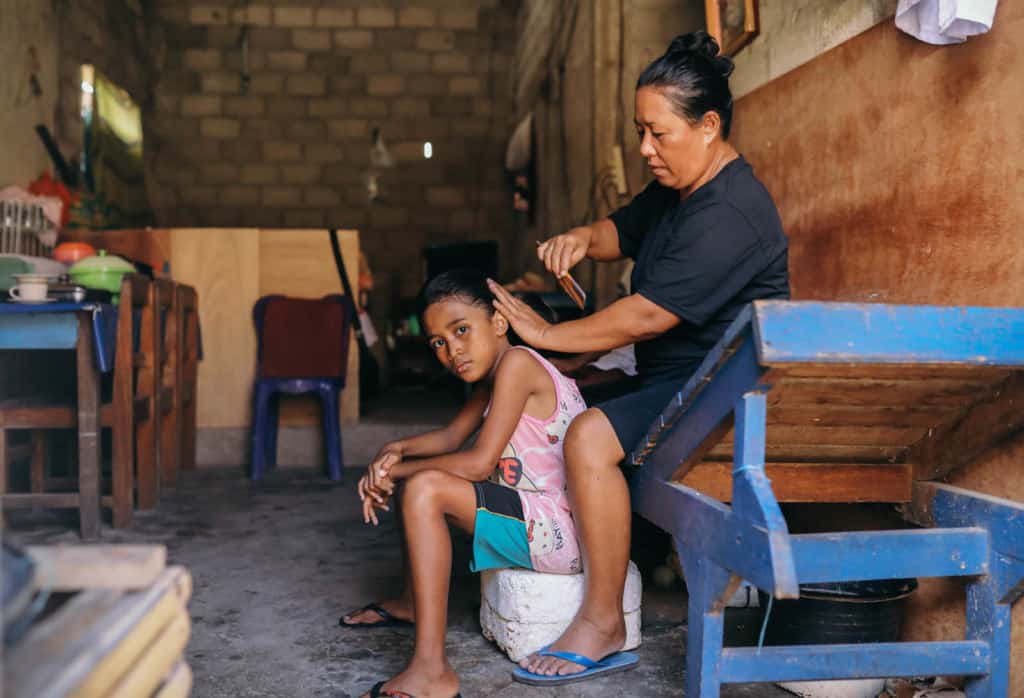 Yosina, wearing a pink and white shirt with blue and black shorts, is sitting down while her mother combs her hair. They are sitting inside their home.