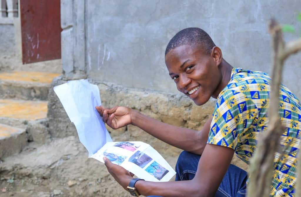Young man wearing a blue and yellow patterned shirt with jeans. He is sitting outside his home and is holding a letter from his sponsor.