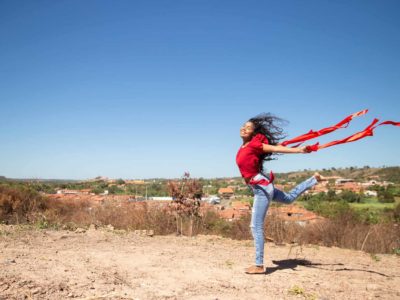 Maria Rita is wearing a red shirt and jeans. She is holding a red ribbon in each hand is jumping in the air. Her town is in the background.