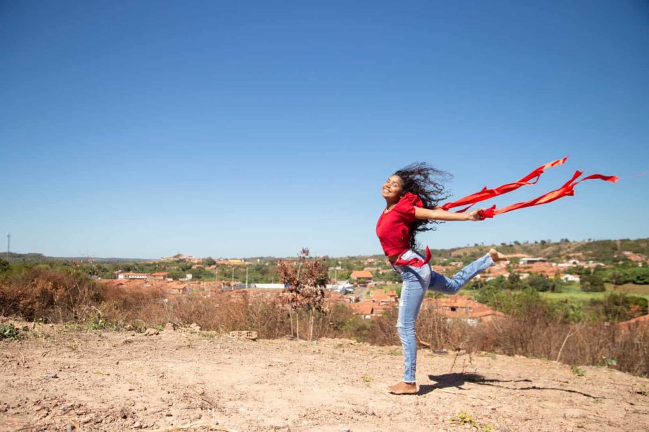 Maria Rita is wearing a red shirt and jeans. She is holding a red ribbon in each hand is jumping in the air. Her town is in the background.