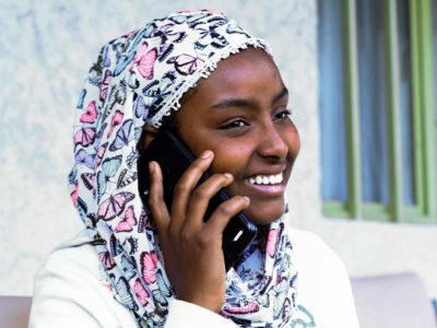 Woman wearing a white shirt and a floral print head covering. She is sitting outside and is talking on the phone.