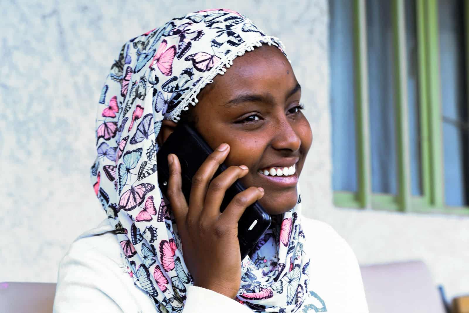 Woman wearing a white shirt and a floral print head covering. She is sitting outside and is talking on the phone.
