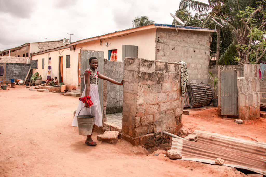 Adrienne is wearing a white skirt with a brown, red, and white shirt. She is wearing a red cloth tied around her wrist and is carrying a bucket into the entrance to her home.