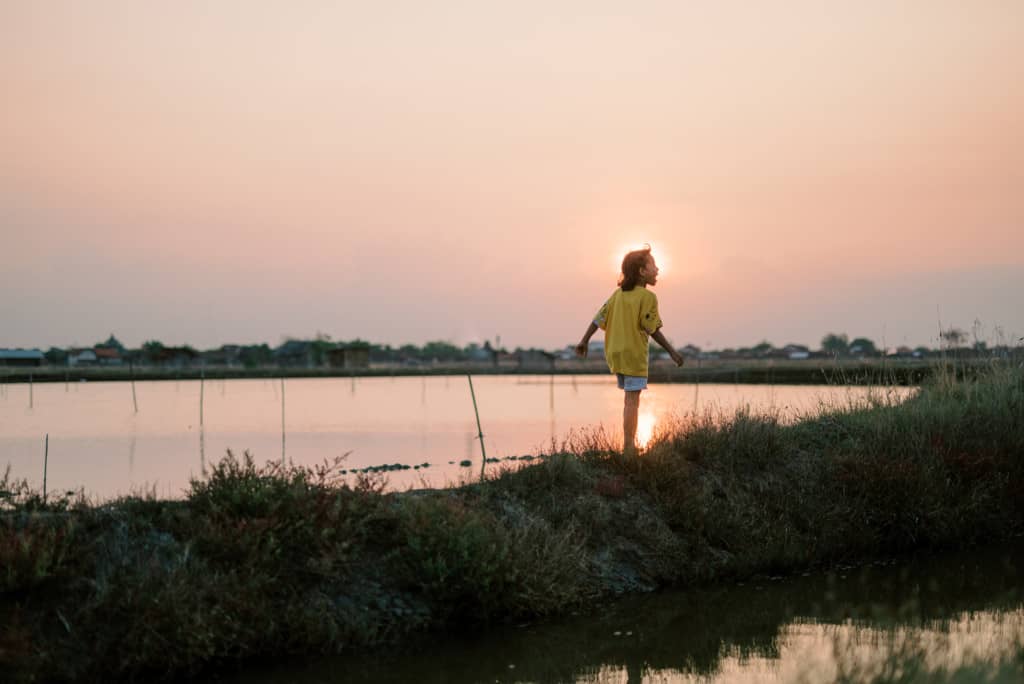 A girl wearing shorts and a yellow shirt walks along the banks of a salt pond in Indonesia. The sun is setting in the distance and reflecting on the water.