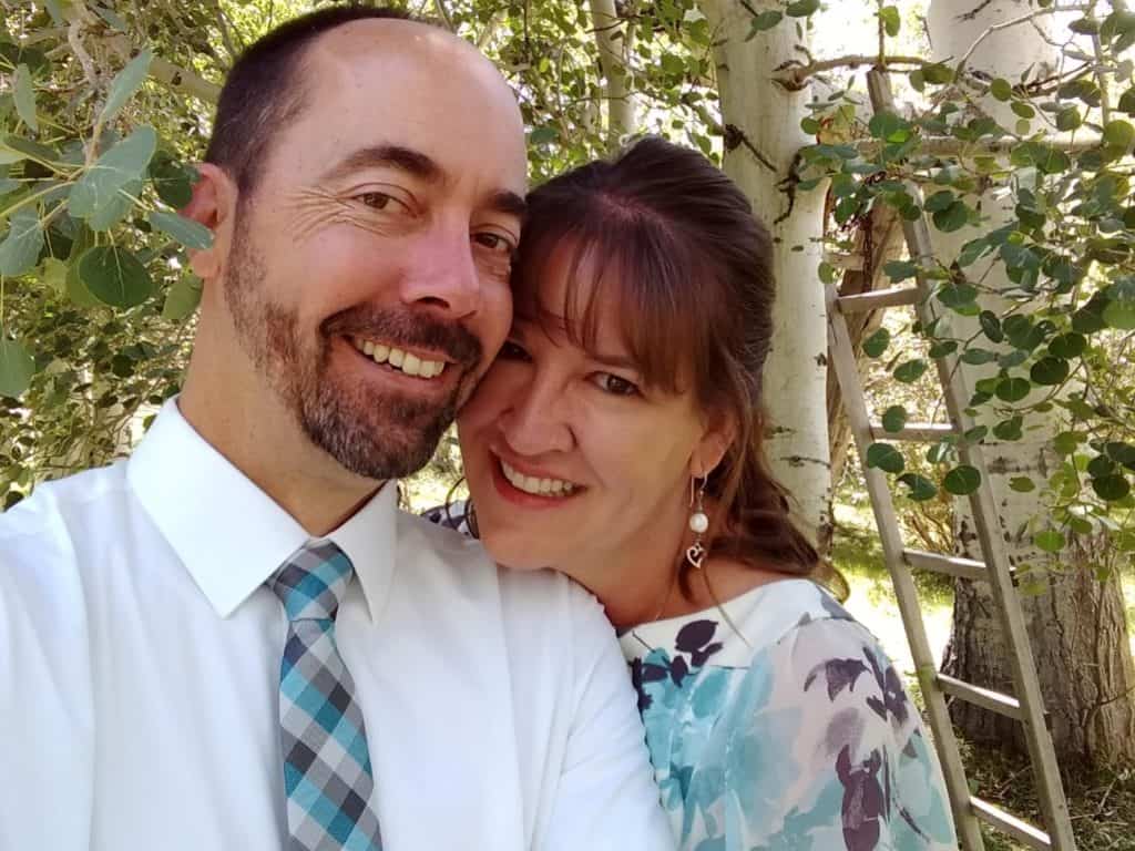 Jeff and Bonnie smile as they stand close to each other. He is wearing a white shirt and blue and gray tie. She is wearing a floral blouse.