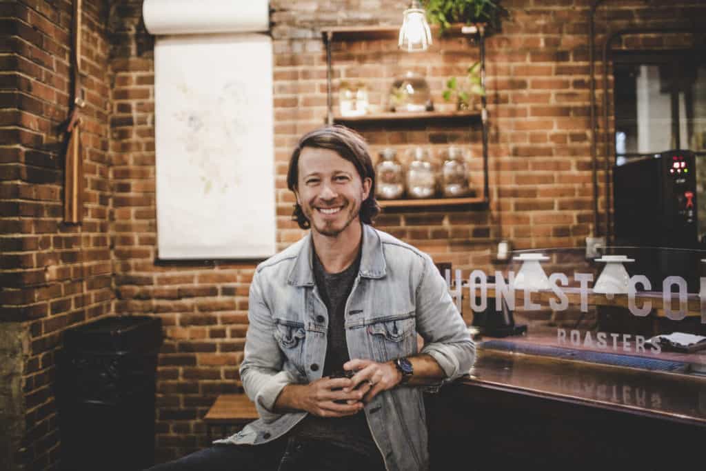 Mike Donehey, wearing a gray T-shirt and denim jacket, sits at a coffee roasting bar. There is a brick wall behind him.