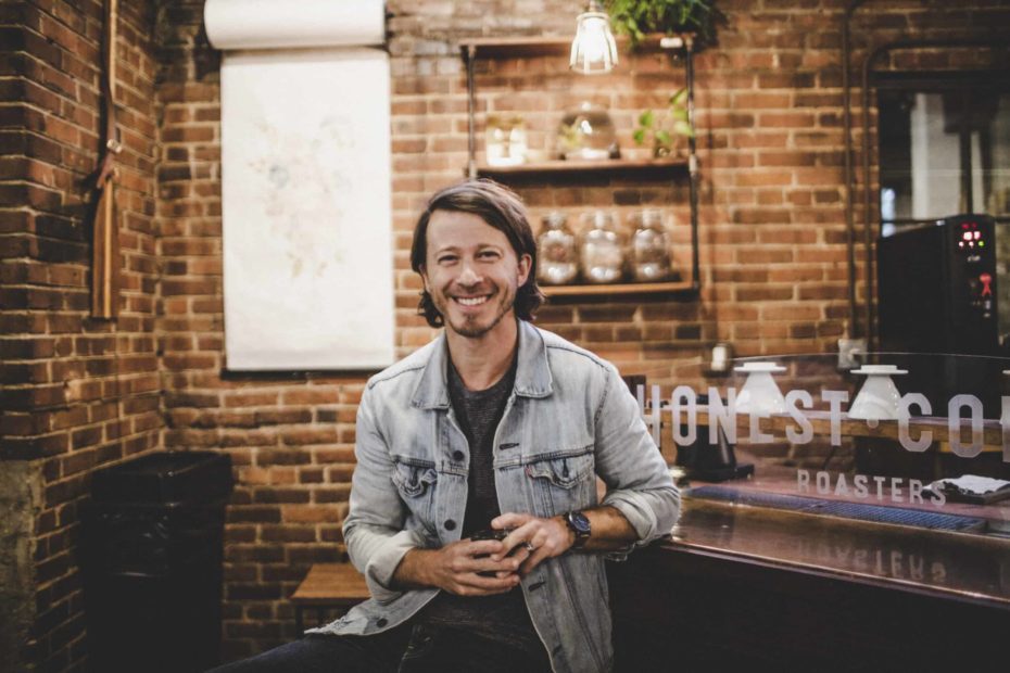 Mike Donehey, wearing a gray t-shirt and denim jacket, sits at a coffee roastery. There is a brick wall behind him.