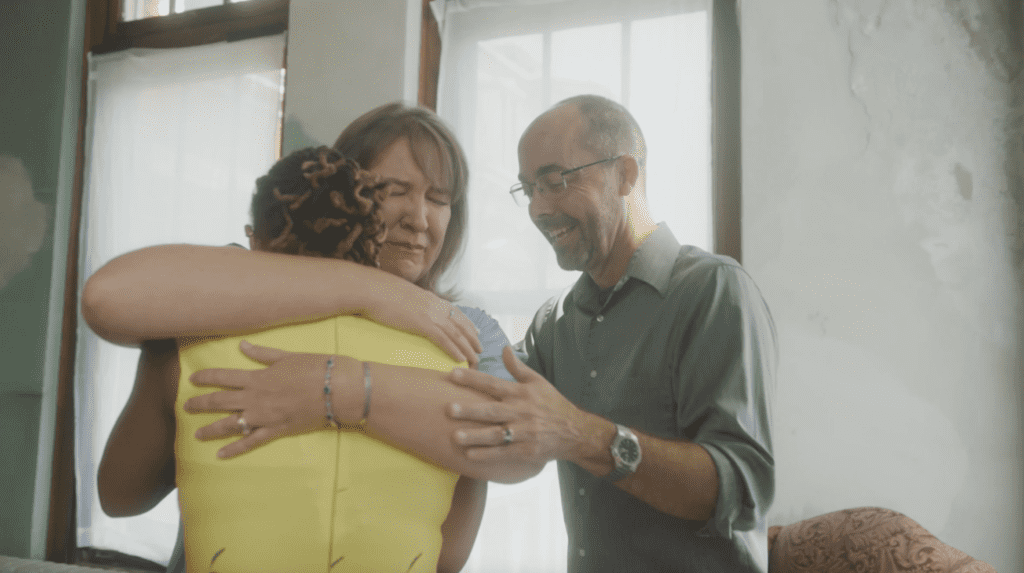 Two people embrace a woman wearing a yellow dress.