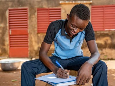 A young man wearing a blue shirt and jeans sits down outside. He is writing a letter.