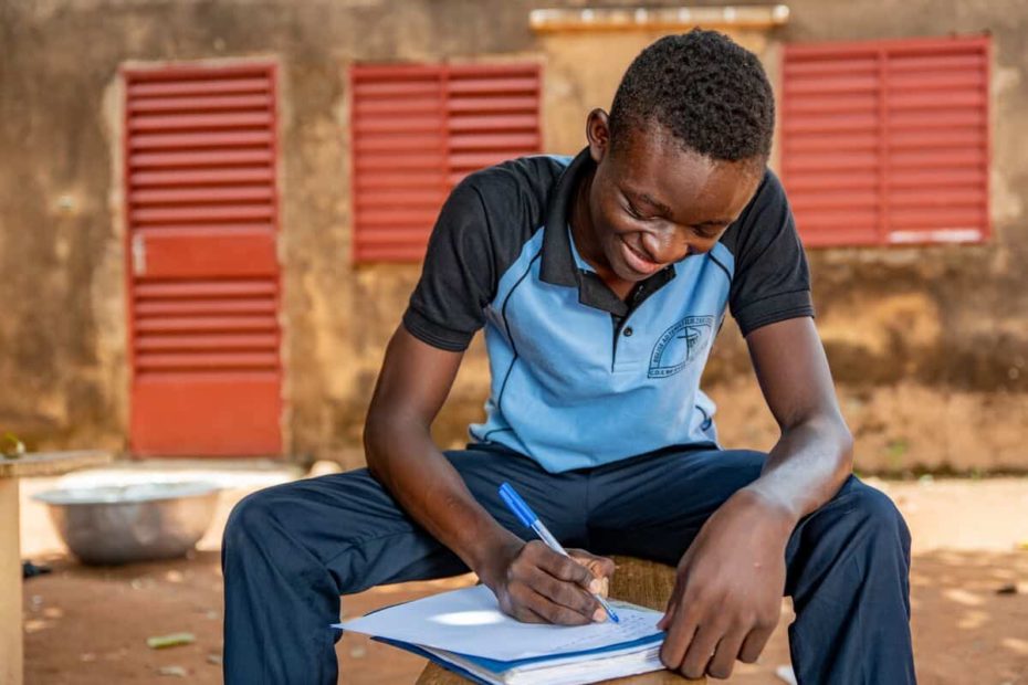 A young man wearing a blue shirt and jeans sits down outside. He is writing a letter.