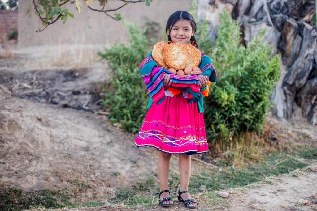 Sheyla is holding a food basket with bread, eggs, and other nutritious food she receives in the food basket the church gives her. She is wearing traditional clothing. 