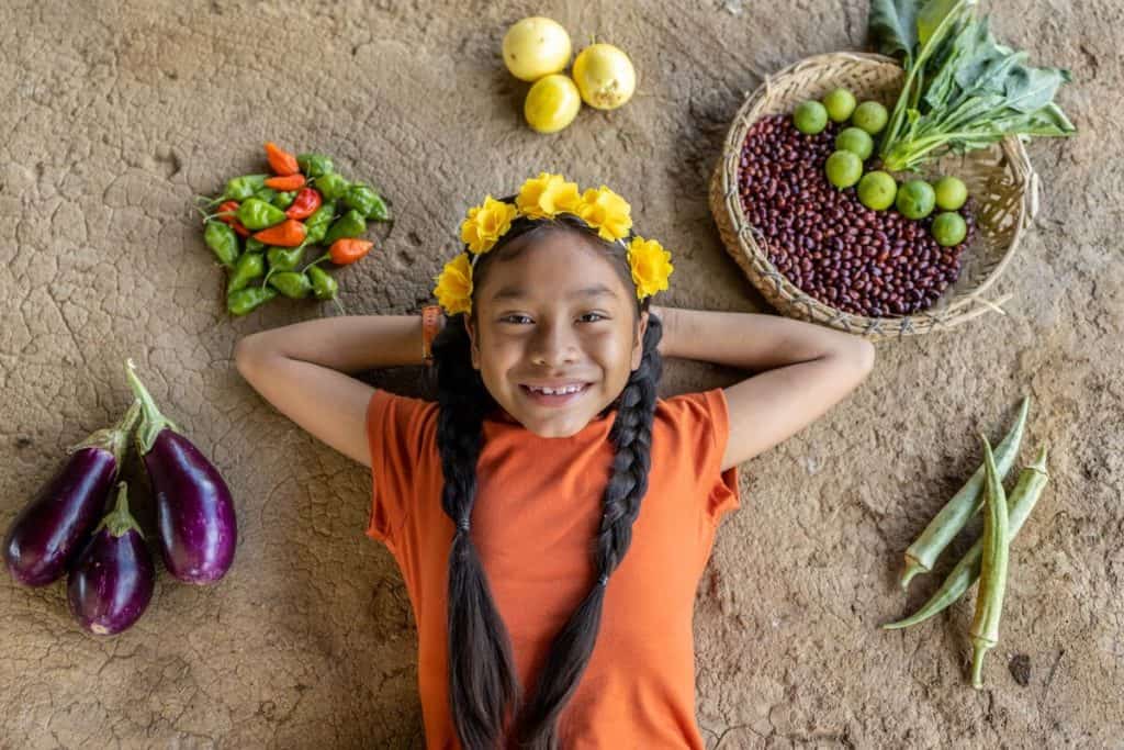 Maira is wearing an orange shirt with jeans. She is laying down at her home and is surrounded by vegetables from her garden.