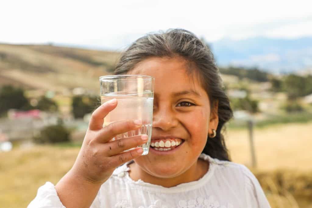 Brittany in Colombia is thankful for clean water. She is wearing a white shirt and an orange skirt. She is standing outside and is holding a glass of clean water in front of her face.