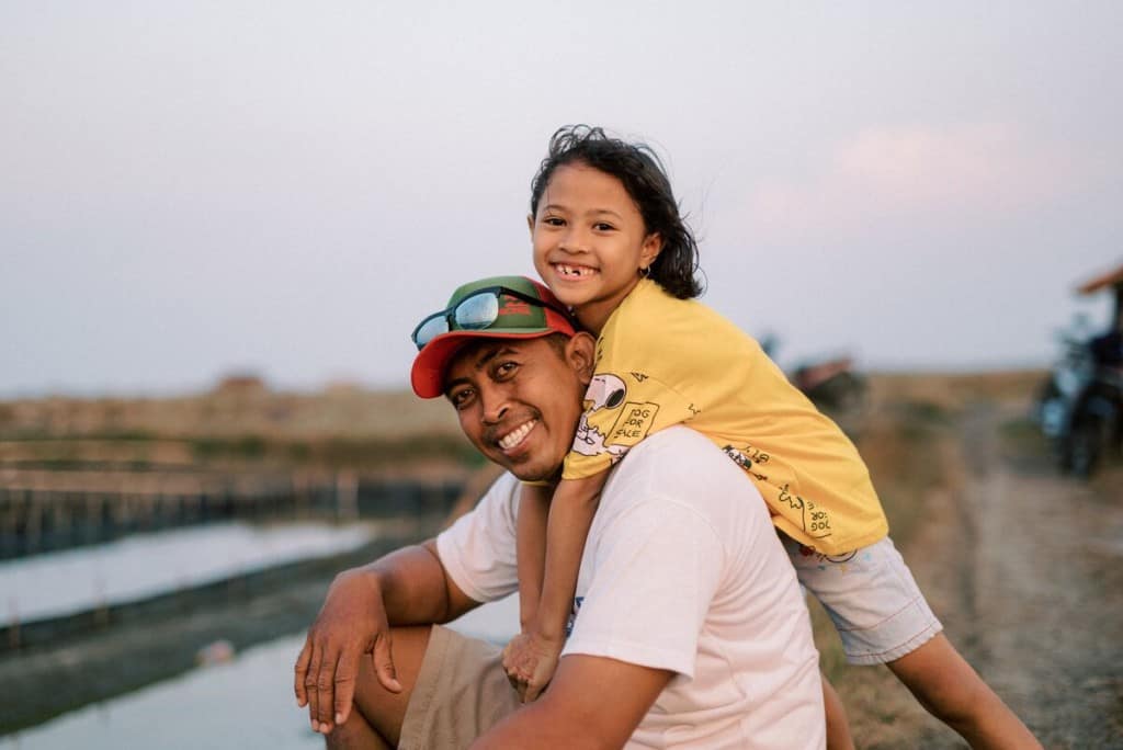 A father and daughter in Indonesia smile. The father is wearing sunglasses on his hat and a white shirt. The girl is wearing shorts and a yellow shirt.
