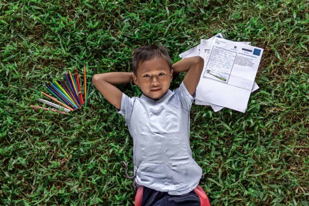 Anllelo is thankful for letters from his sponsor. He wearing a gray shirt. He is laying down in the grass with his hands behind his head. On one side of him are some of his sponsor's letters. On the other side are colored pencils. 