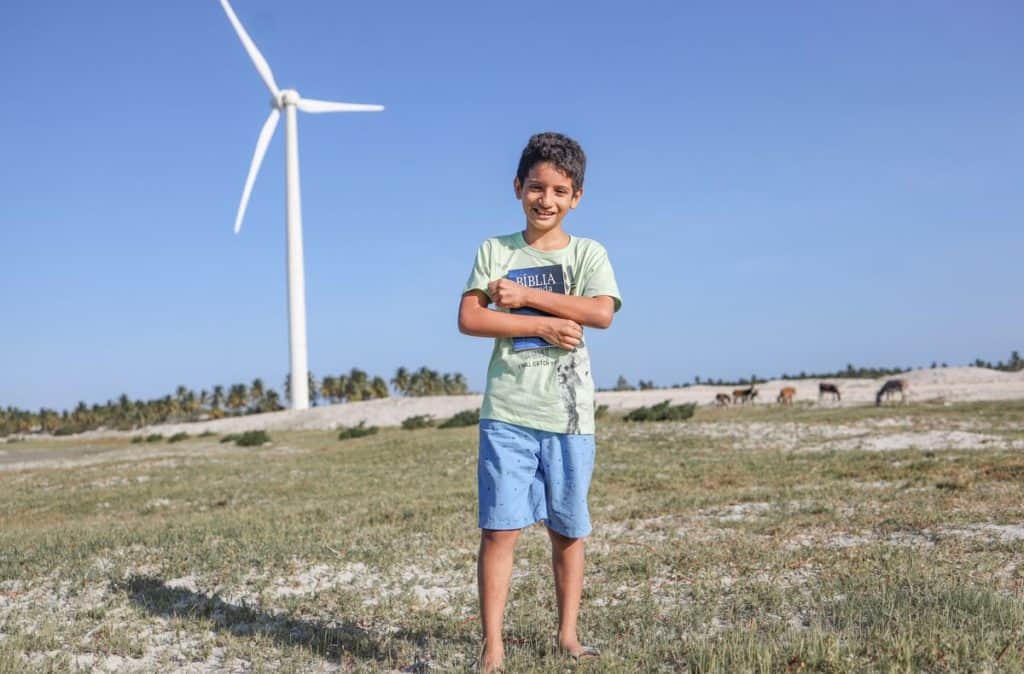 Iarley is wearing a green shirt and blue shorts. He is standing outside and is holding a Bible close to his chest. There is a windmill in the background. 