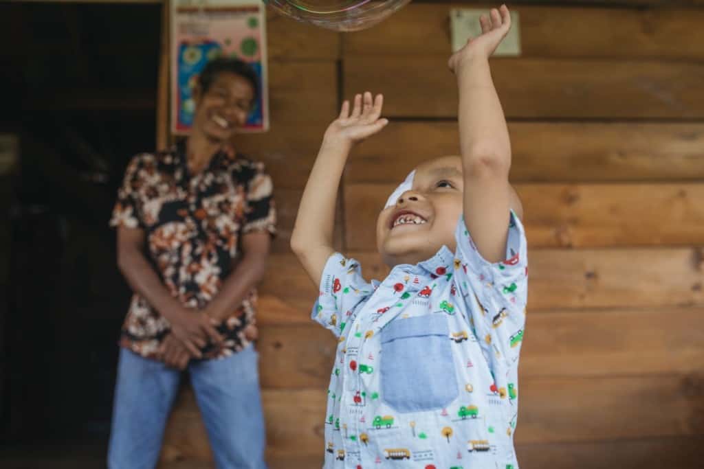 Yoskiel is wearing a light blue shirt with cars, trucks, and trains on it. He has an eye patch. Yoskiel is standing on the front porch of his home and is looking up at balloons he is playing with. His father is in the background.