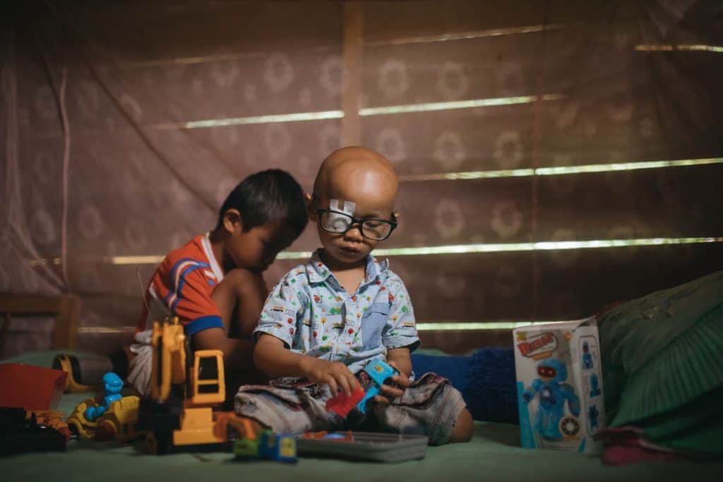 Yoskiel is wearing a light blue shirt with cars, trucks, and trains on it. He has an eye patch and is wearing glasses over it. He is sitting on the floor of his home and is playing with his brother.