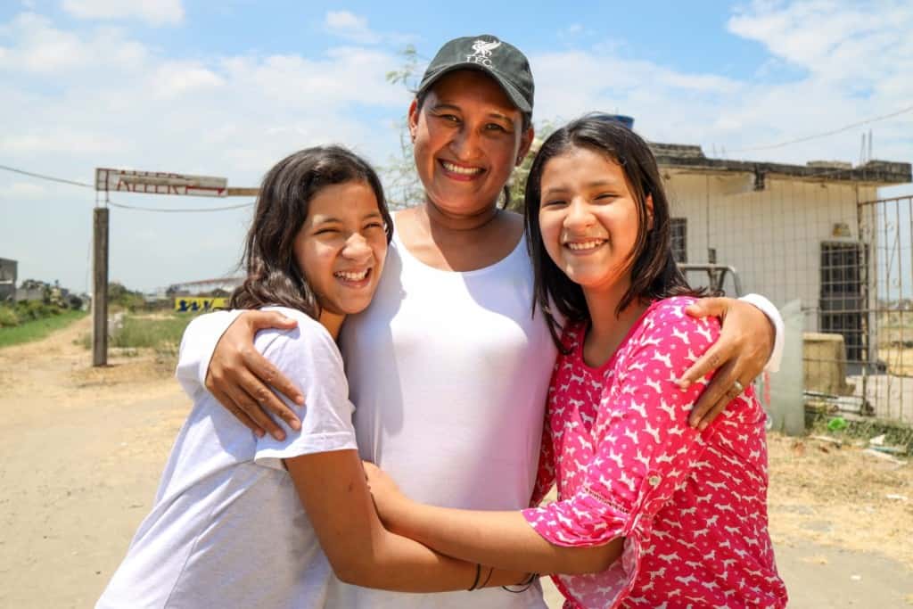 Cintya is wearing a white shirt and jeans. Joselen is wearing a pink shirt and jeans. They are standing outside the church and are hugging their tutor, Jenny, wearing a white shirt and a hat.