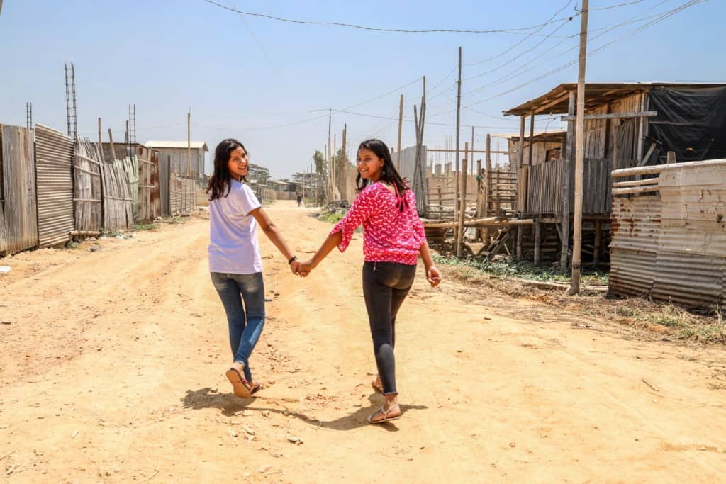 Cintya is wearing a white shirt and jeans. Joselen is wearing a pink shirt and jeans. They are holding hands and walking down a dirt road in their neighborhood. They are walking away from the camera but looking back.