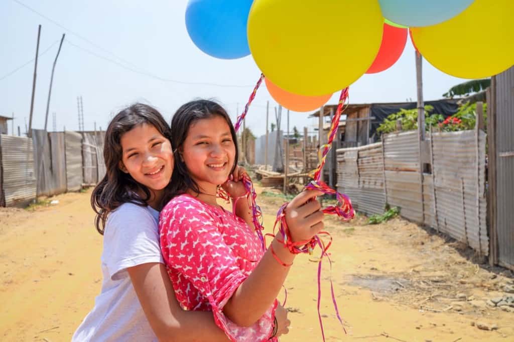 Cintya is wearing a white shirt and jeans. Joselen is wearing a pink shirt and jeans. They are both holding colorful balloons and are playing in their neighborhood.