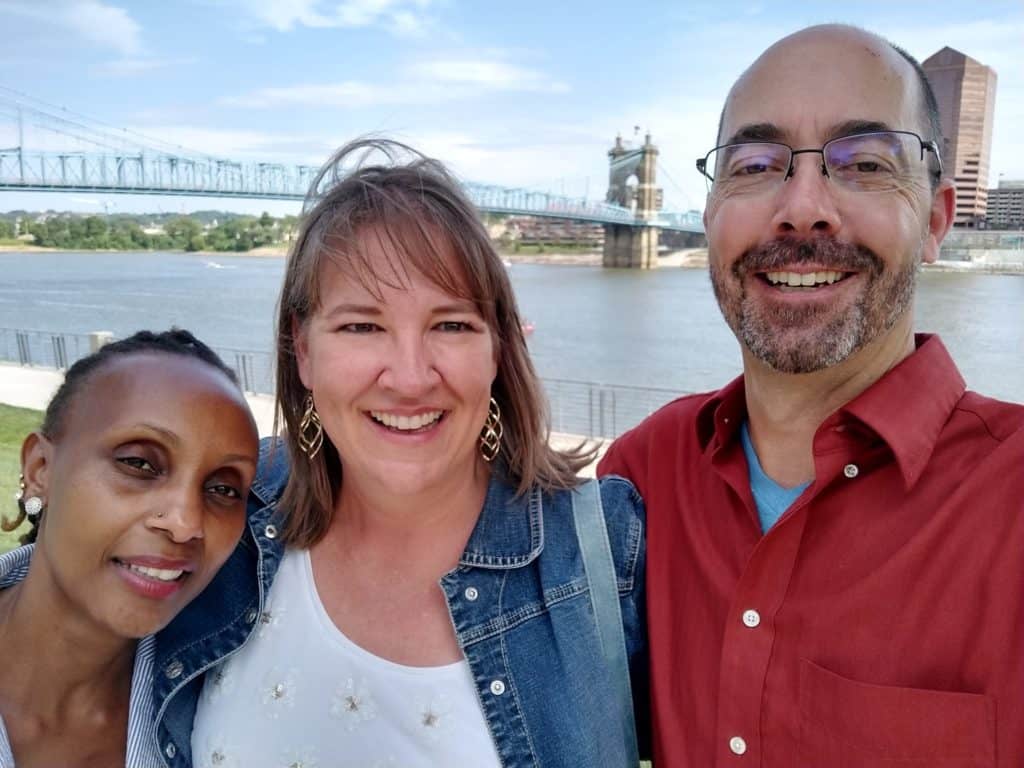 Two women and a man stand together, smiling. There is a bridge and water in the background.