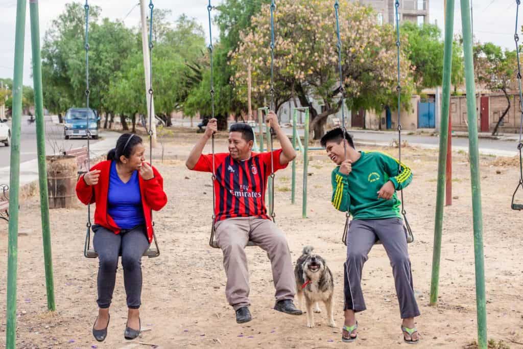 A father and his two children, teenagers, swing on a green swing set at a playground in Bolivia. A dog is with them.