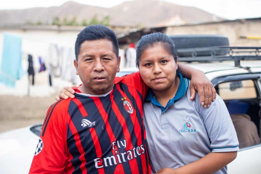 A father puts his arm around his teenage daughter. He is wearing a Fly Emirates soccer jersey, and she is wearing a polo shirt.