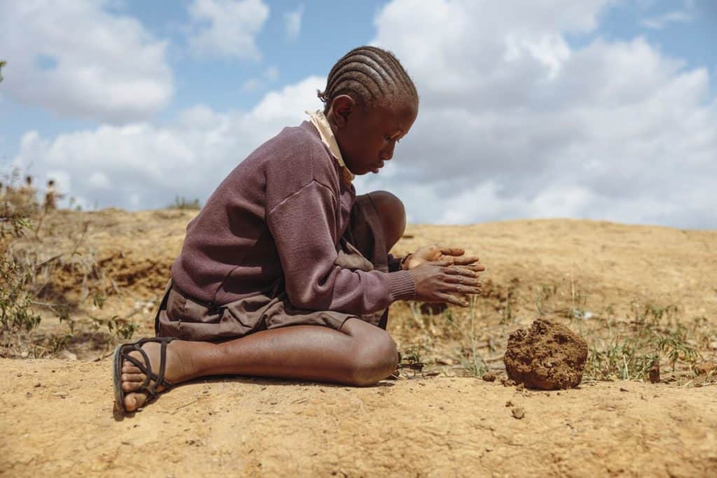 Neema is wearing a brown sweater and brown skirt. She is sitting down outside her school and is using clay to make a figurine.