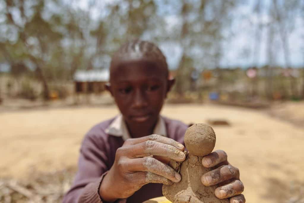 Neema is wearing a brown sweater and brown skirt. She is sitting down outside her school and is making something out of clay. She is sculpting on a small yellow barrel.