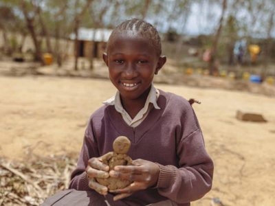 Neema is wearing a brown sweater and brown skirt. She is sitting down outside her school and is holding a figurine she sculpted.