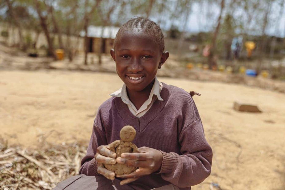 Neema is wearing a brown sweater and brown skirt. She is sitting down outside her school and is holding a figurine she sculpted.