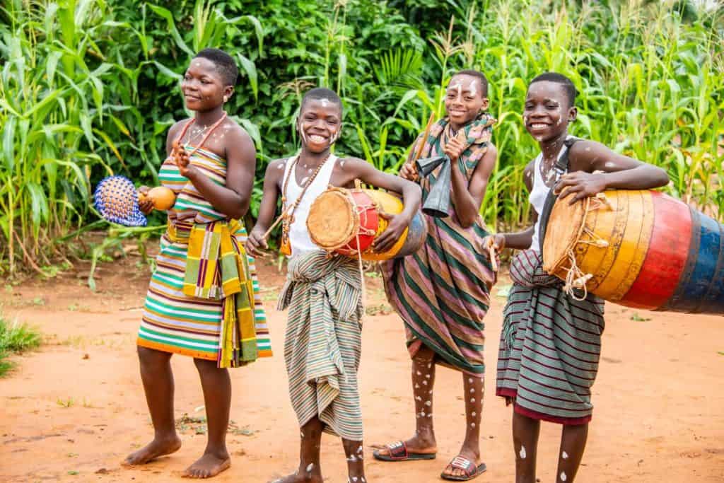Four children, youths, are standing outside playing musical instruments and dancing. There are trees and tall grass behind them. The children are wearing traditional clothing of Togo.