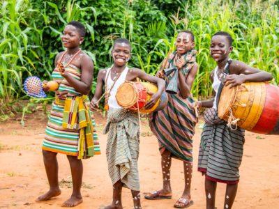 Four children are standing outside playing musical instruments and dancing. There are trees and tall grass behind them. The children are wearing traditional clothing.