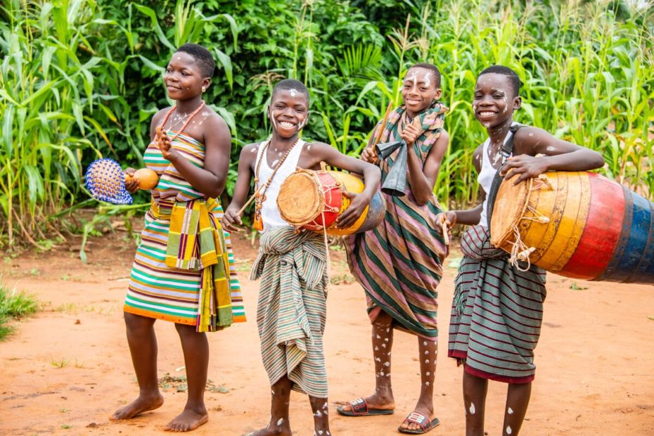 Four children are standing outside playing musical instruments and dancing. There are trees and tall grass behind them. The children are wearing traditional clothing.