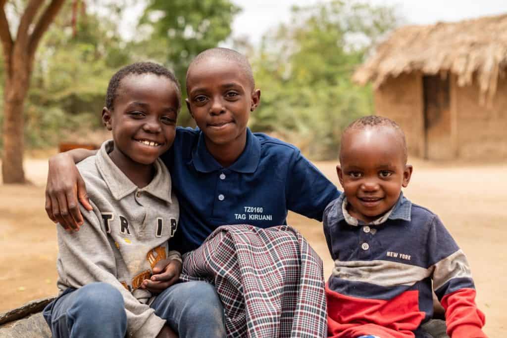 Three children in Tanzania sit together smiling. A mud hut with thatch roof is in the background.