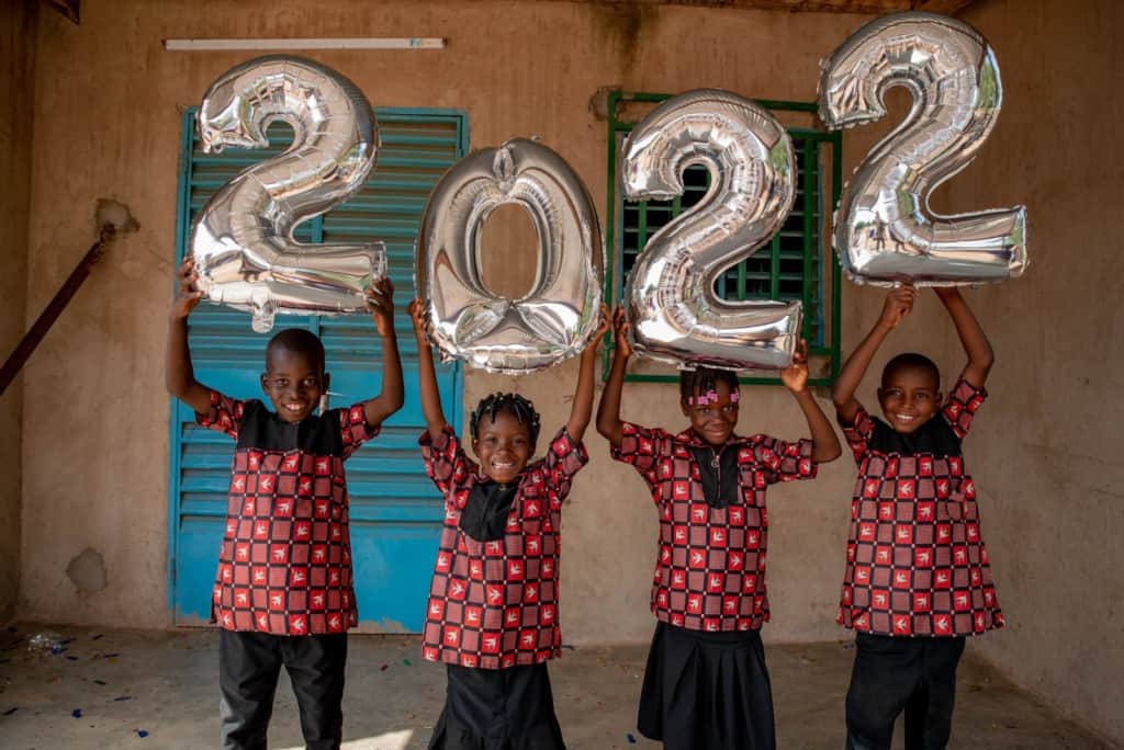 A group of children are together outside the center. They are wearing red and black patterned shirts with black pants or a black skirt. Moussa, Christelle, Milka, and Alfred are holding the numbers 2022 made with silver balloons.