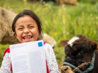 Lizbeth is wearing a blue skirt, white shirt, and a red sash. She is sitting in a field with a calf behind her and she is holding up one of her sponsor's letters.