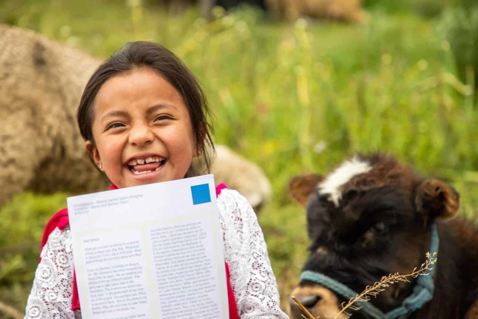 Lizbeth is wearing a blue skirt, white shirt, and a red sash. She is sitting in a field with a calf behind her and she is holding up one of her sponsor's letters.