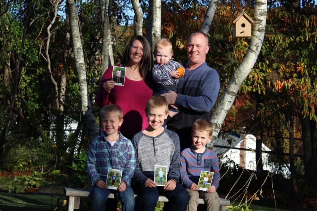 A man and woman stand outside with their four sons. The man is holding a toddler, and three boys are sitting on a bench holding photos of children their family sponsors. There are trees and a bird feeder behind them.